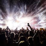 A crowd of people at a concert holds up phones and hands as bright stage lights and confetti fill the air.