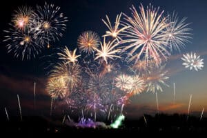 A large display of colorful fireworks explodes in the night sky above a dark landscape, with bright bursts and trails lighting up the scene.