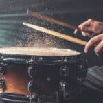Close-up of a person striking a snare drum with drumsticks, causing particles to rise from the drumhead in dramatic lighting.