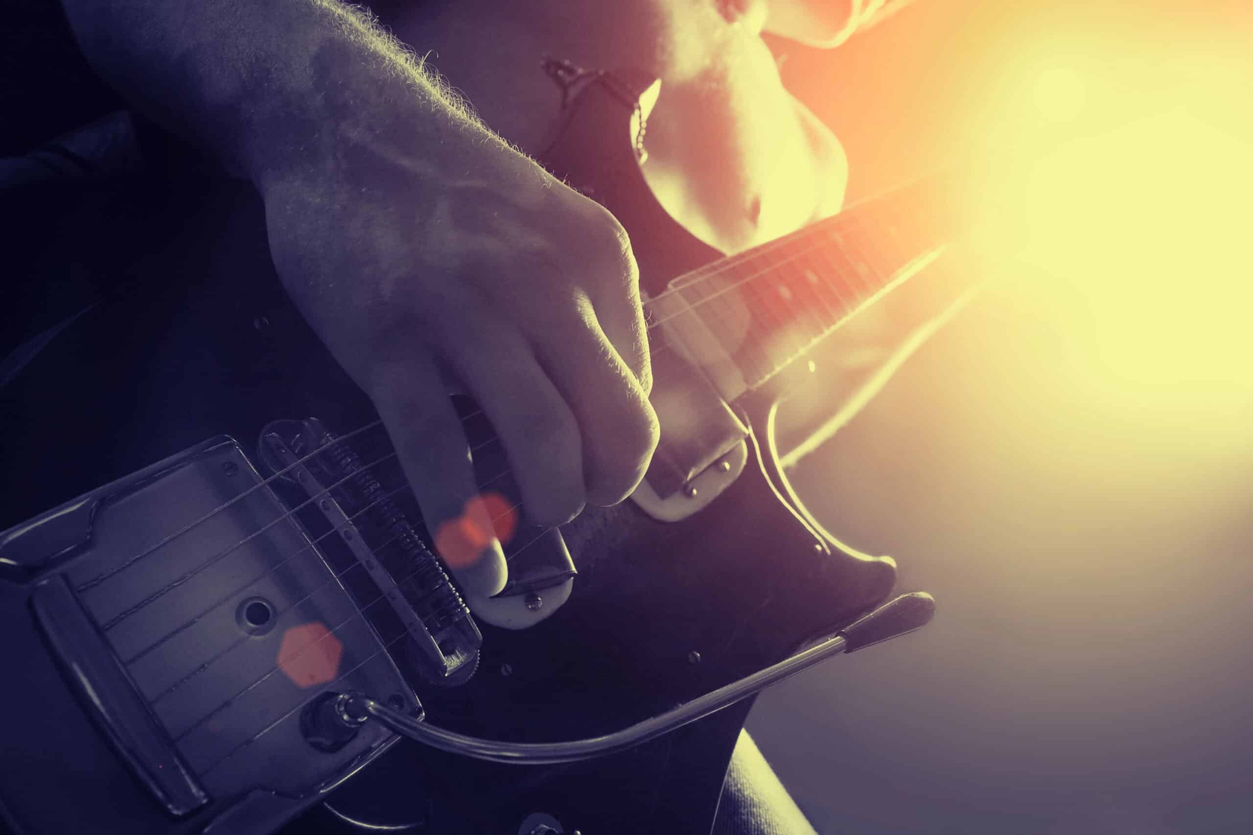 Close-up of a person playing an electric guitar, with sunlight and lens flare illuminating the scene from the right.