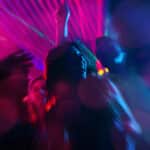 A group of people dance under colorful neon lights at a party, with blurred motion and vibrant pink and blue hues creating a dynamic atmosphere.