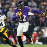 A football player in a purple Ravens uniform runs with the ball while fending off two defenders in white Steelers uniforms during a game.