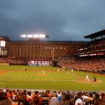 A baseball game in progress at a stadium with fans in the stands, illuminated scoreboard, and players on the field under an overcast sky.