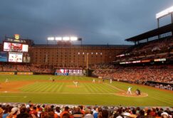 A baseball game in progress at a stadium with fans in the stands, illuminated scoreboard, and players on the field under an overcast sky.