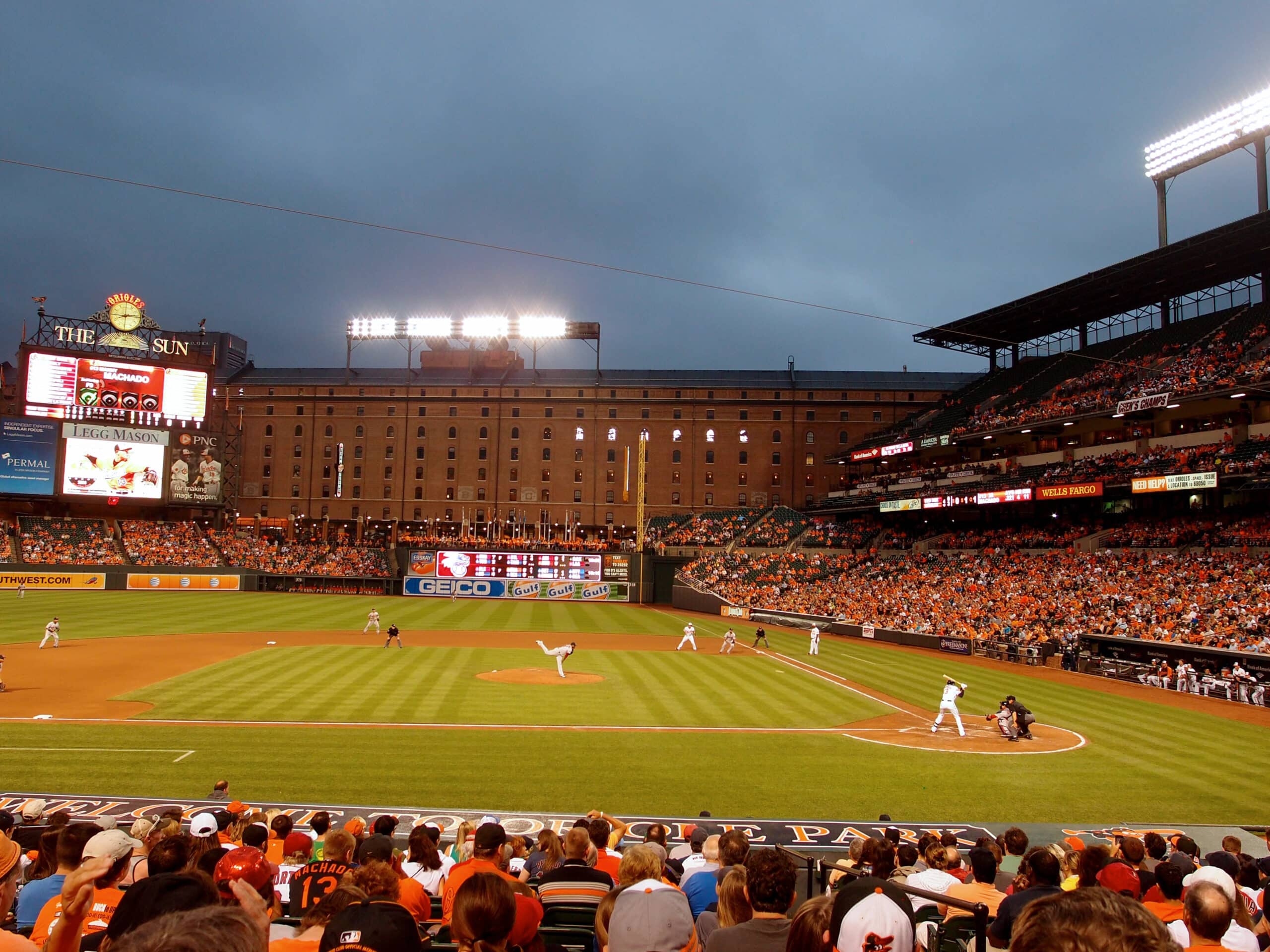 A baseball game in progress at a stadium with fans in the stands, illuminated scoreboard, and players on the field under an overcast sky.