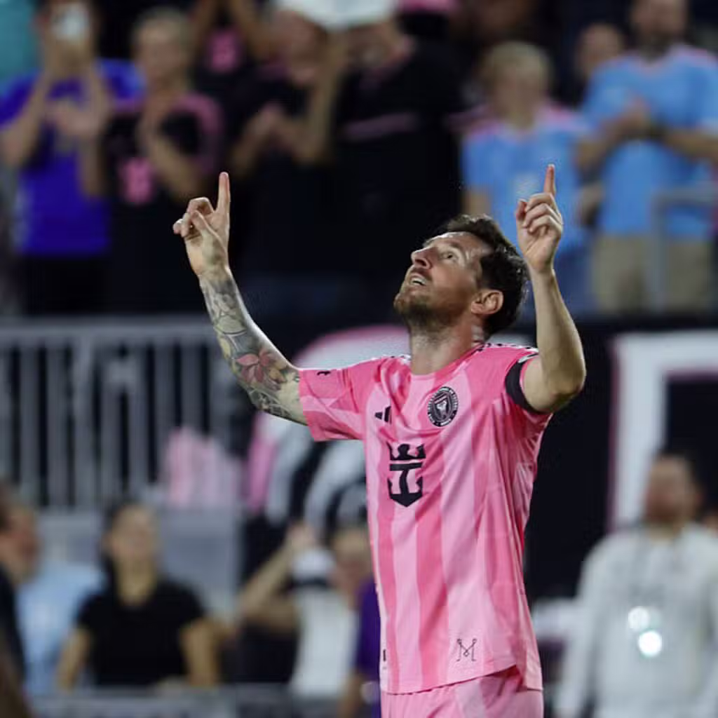 A soccer player in a pink and black uniform raises both hands and points upwards on a field, with a blurred crowd in the background.