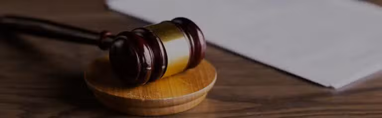 A wooden judge's gavel rests on a sound block beside a white sheet of paper on a wooden surface.