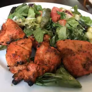 Four pieces of grilled tandoori chicken served with a side of mixed green salad on a white plate.