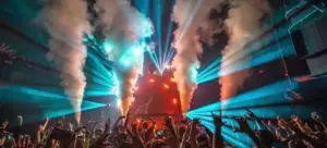 A crowd at a concert or nightclub raises their hands as colorful stage lights and smoke effects illuminate the scene.